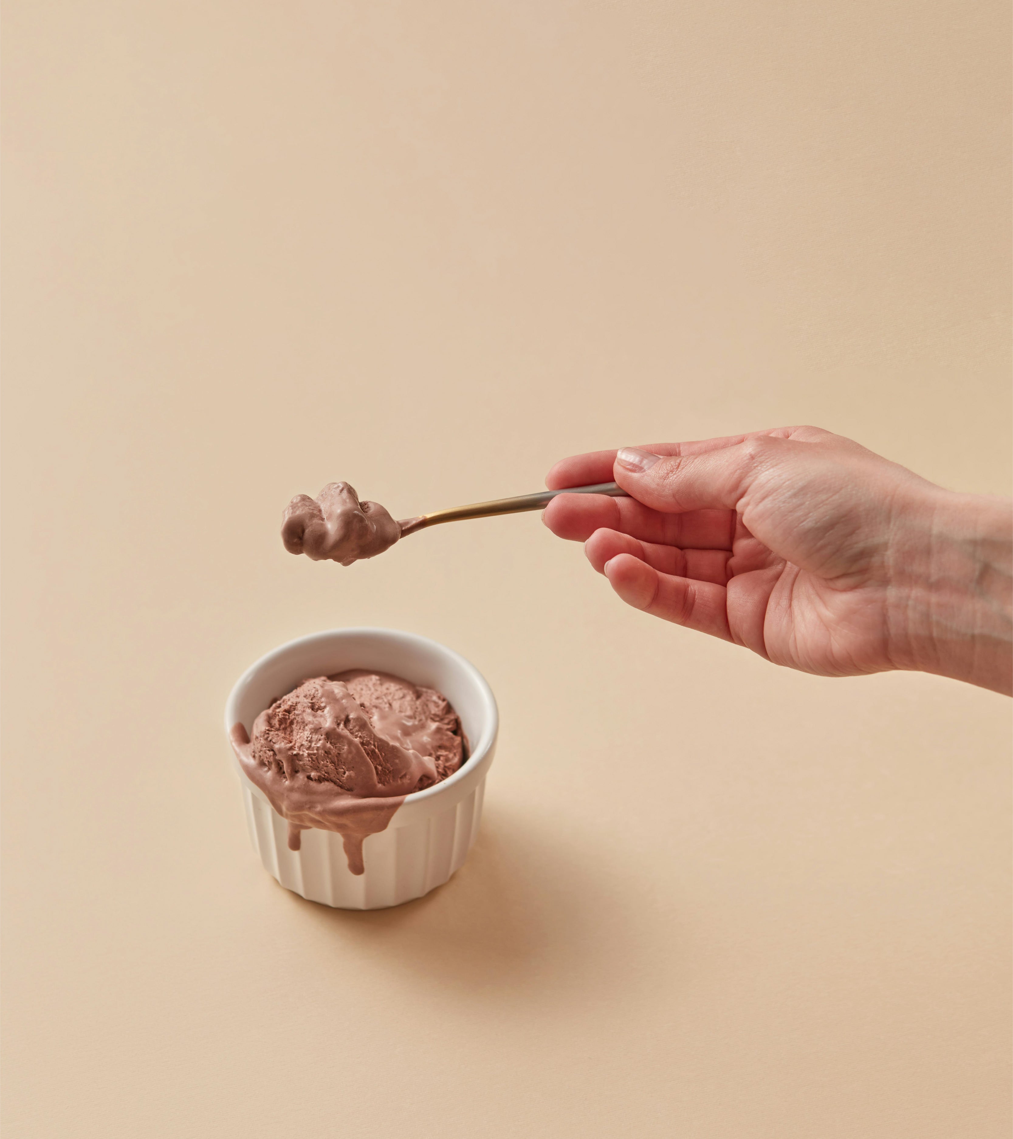 Close up of a woman's hand grabbing a bronze spoon with chocolate ice cream. Below is a white ramekin filled with chocolate ice cream. Dark beige color background