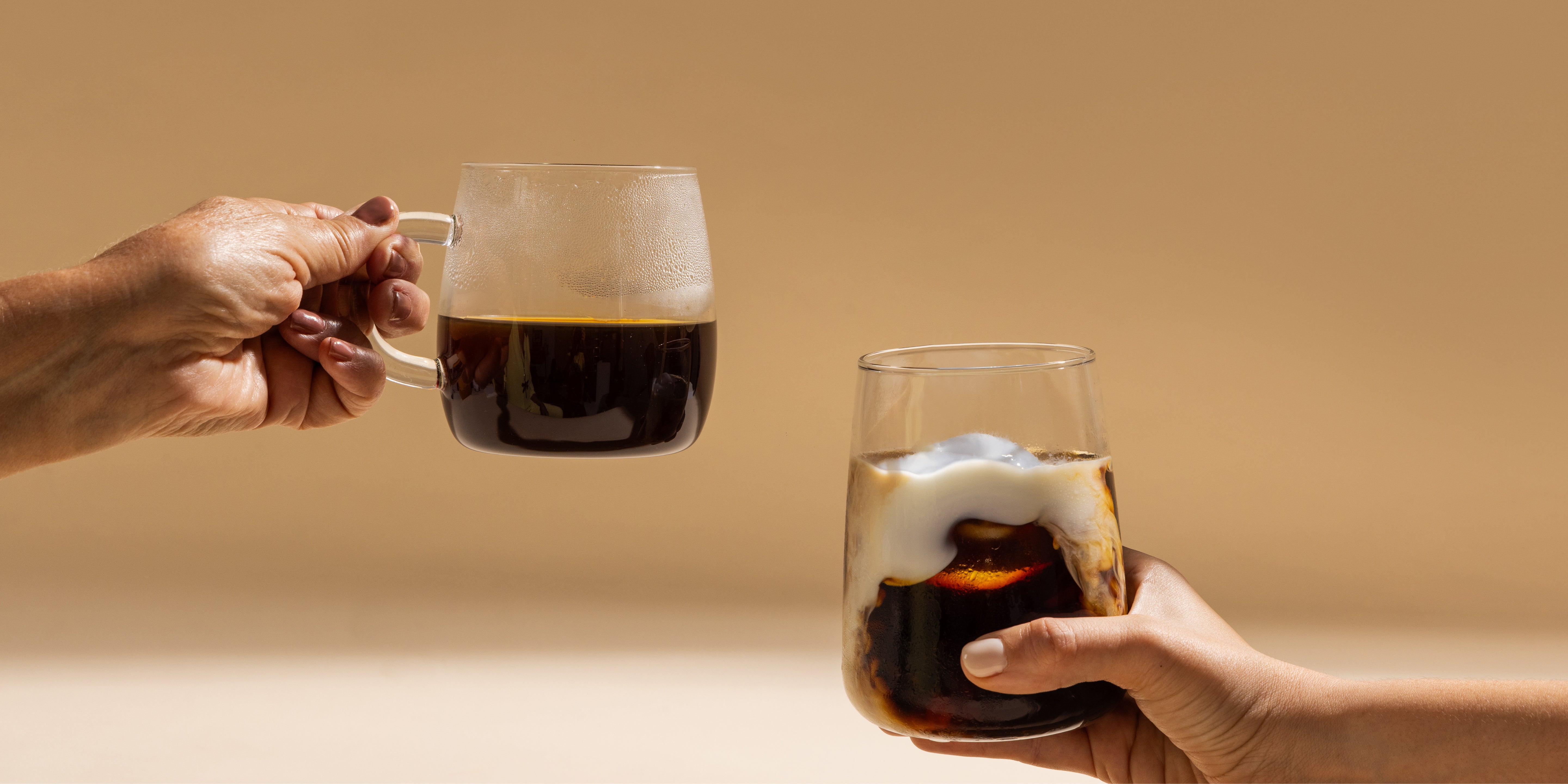 Two female hands , one is older than the other grabbing clear coffee glasses. One hot, one cold.